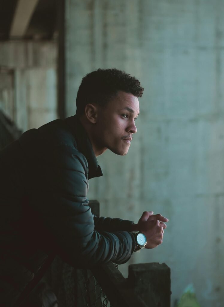 Young adult man leaning on metal railing looking pensive, wearing casual attire.